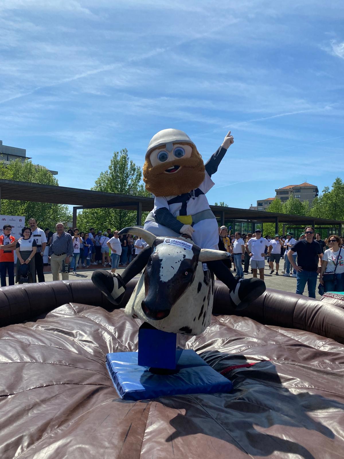 Mascote de um evento a andar no touro mecânico, com público à volta